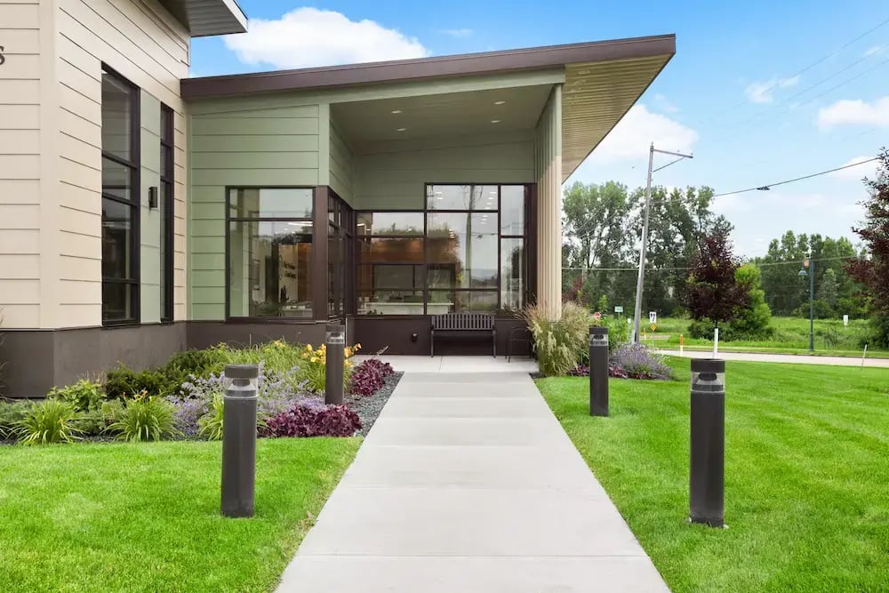 Concrete walkway to modern endodontic office entrance with sage green siding, large black-framed windows, covered portico, and landscaped grounds with ornamental plants and bollard lighting