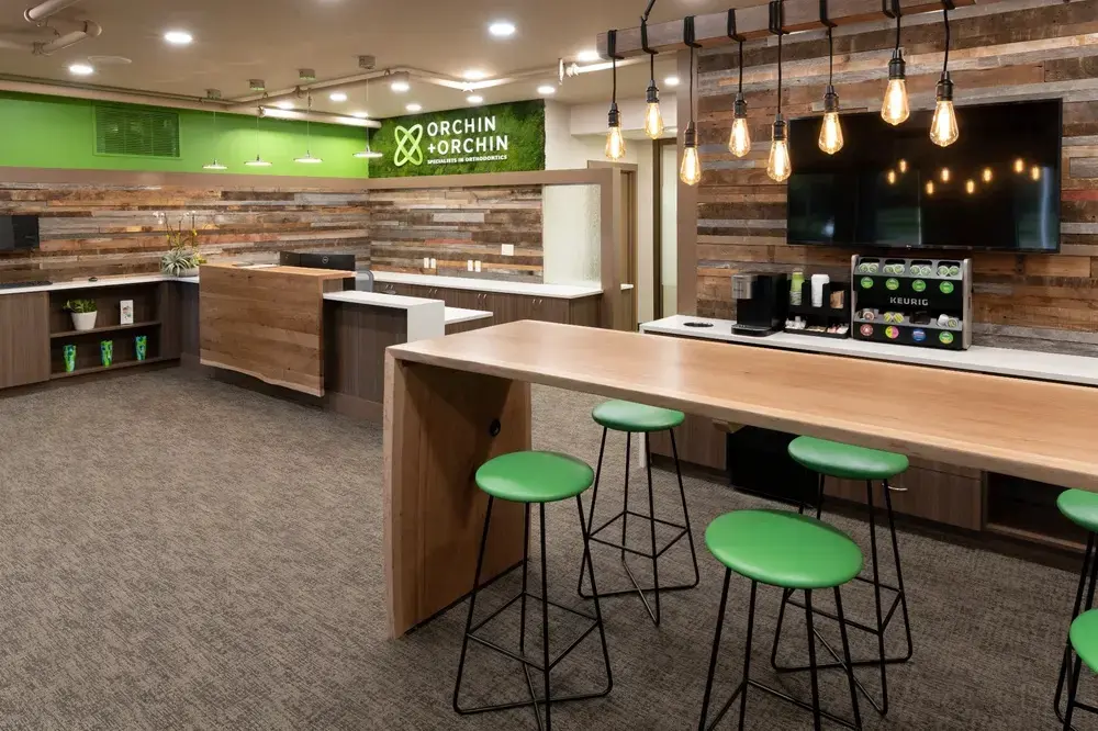 Contemporary Orchin + Orchin Orthodontics reception and waiting area featuring reclaimed wood accent walls, green brand elements including reception desk backsplash, light wood countertop with green bar stools, Edison bulb pendant lighting, and modern coffee station