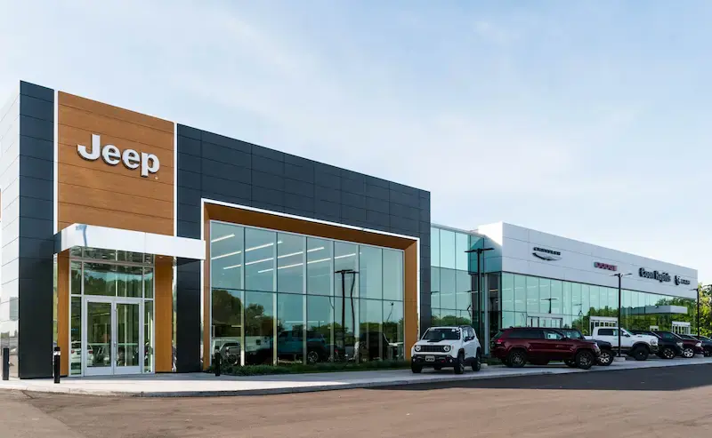 Exterior of a modern Jeep Chrysler Dodge Ram dealership with dark metal and wood-accented facade, large glass showroom windows, and SUVs parked on the front lot under a clear blue sky.