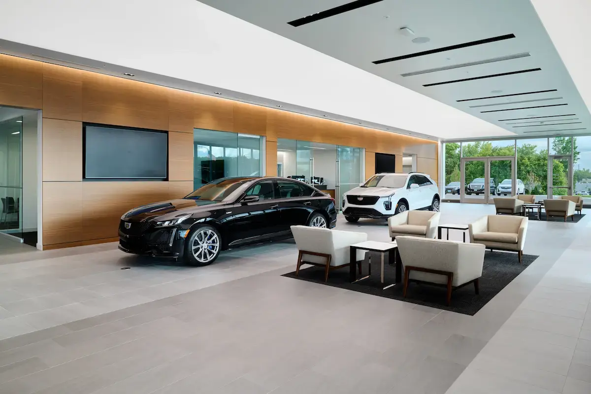 Modern Cadillac showroom featuring a black CT5-V sedan and white XT4 SUV on display, with warm wood-paneled walls, recessed lighting, and a customer lounge area with cream armchairs. Floor-to-ceiling windows reveal the lot beyond.