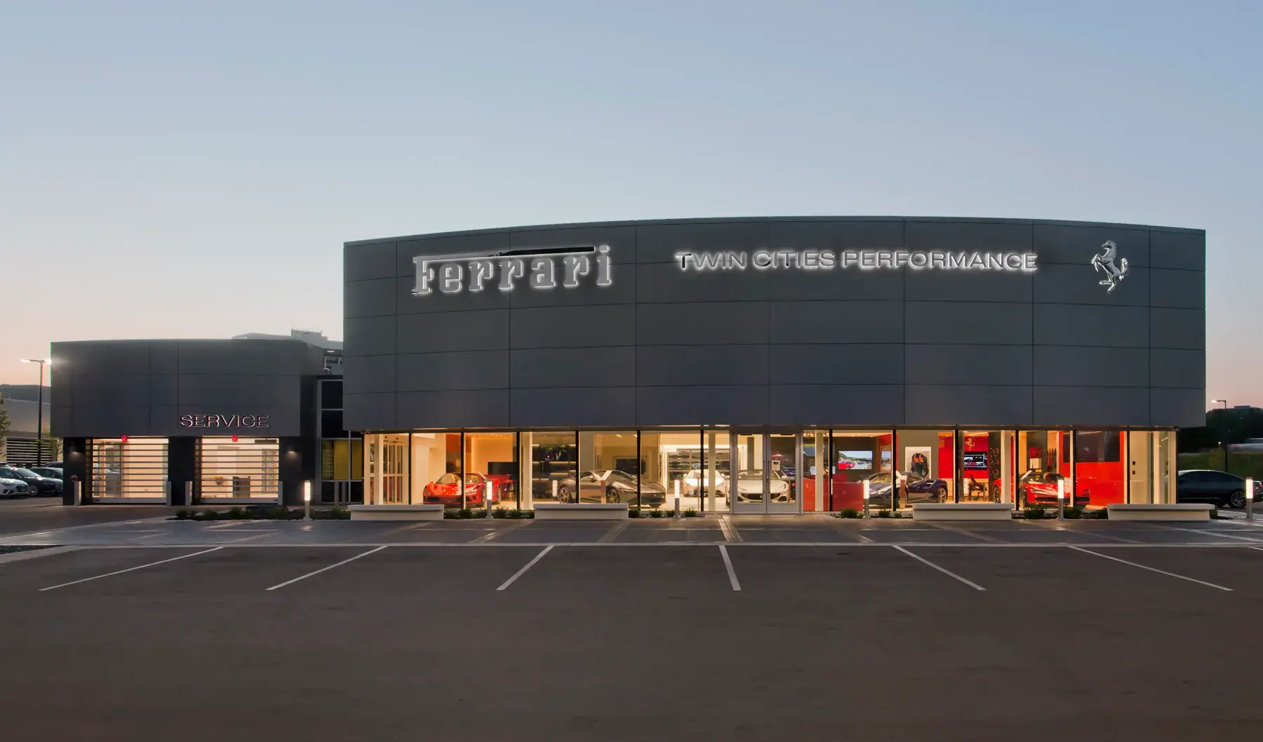 Ferrari Twin Cities Performance dealership at dusk, featuring a curved dark gray facade with illuminated signage and floor-to-ceiling showroom windows displaying red Ferraris inside.