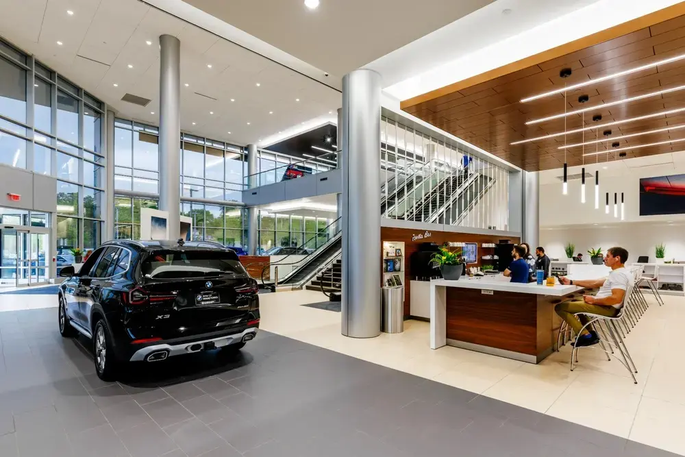 Upscale BMW dealership showroom interior featuring black BMW X3 SUV on display, two-story space with modern staircase, floor-to-ceiling windows, wood-paneled ceiling with linear lighting, Barista Bar cafe area with customers, and contemporary design aesthetic