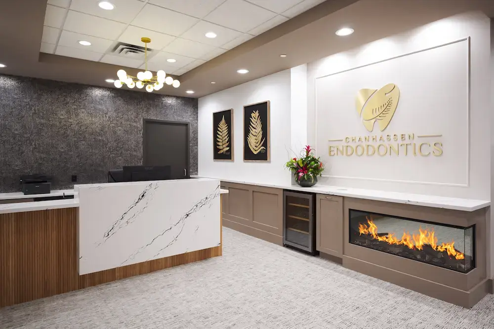 Elegant endodontic office reception area with white marble-look reception desk featuring wood paneling accents, textured dark gray accent wall, gold and white Chanhassen Endodontics branded signage with leaf logo, built-in linear fireplace with taupe cabinetry and gold modern chandelier
