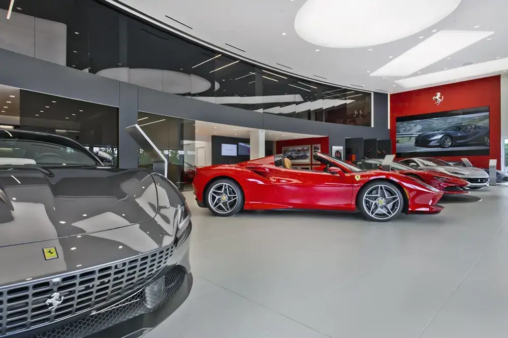 Luxury Ferrari dealership showroom interior featuring red convertible sports car in center with gray Ferrari in foreground, modern ceiling with circular lighting elements, red accent wall with Ferrari prancing horse logo and display screen
