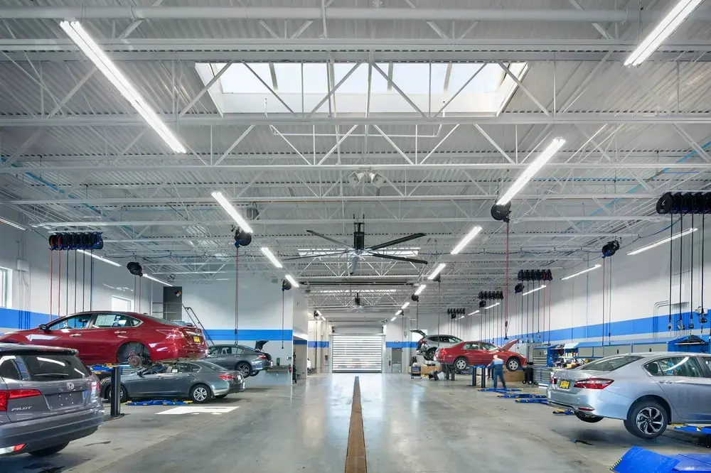 Spacious Honda automotive service bay interior with exposed white metal truss ceiling, multiple vehicle lifts, cars on hoists including red and gray Honda vehicles, bright LED lighting, blue accent stripe along walls, and organized technician workstations