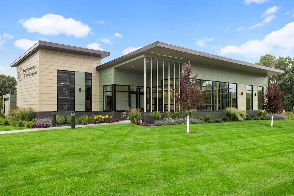 Modern single-story endodontic office building with contemporary architecture featuring sage green and cream horizontal siding, large black-framed windows, covered entryway with vertical slats, and professional landscaping with ornamental grasses and flowering plants on manicured lawn