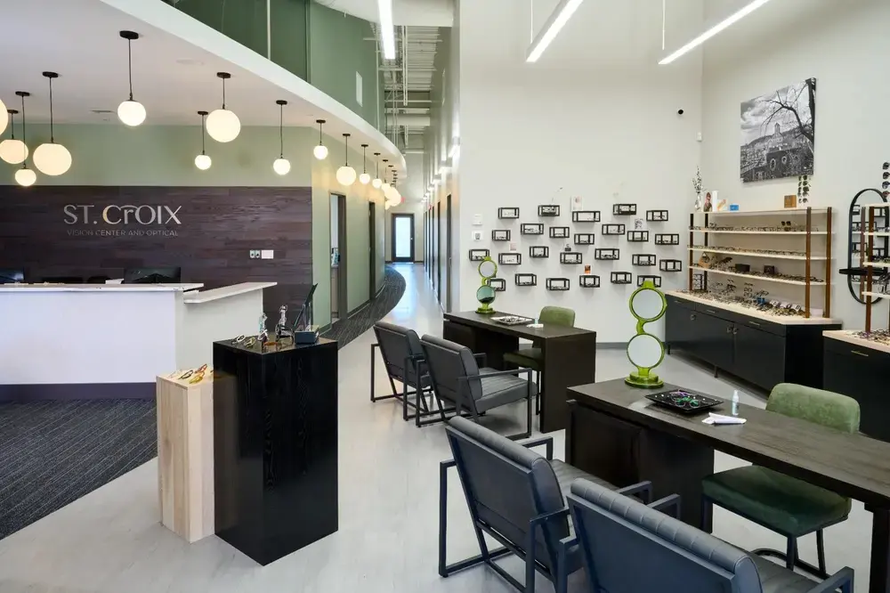 Modern St. Croix Vision Center and Optical retail interior with wooden reception desk, white walls displaying eyeglass frames, green pendant lights, globe pendant lighting, consultation stations with black and green seating, and wood-tone display shelving