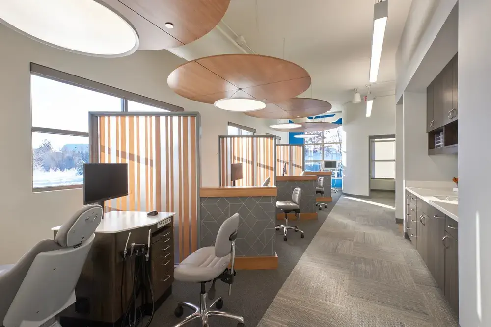 TC Orthodontics treatment room with modern clinical design featuring circular wood acoustic ceiling panels, dental treatment chair and equipment, wood slat privacy dividers with orange and white pattern, large windows with natural light, gray cabinetry, and clean contemporary finishes